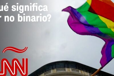 Rainbow pride flag waving beside a Spanish news headline about genero binario above a city building.