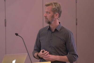 Presenter at a podium giving a lecture about genetisk modificering with a laptop and headset microphone
