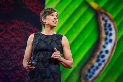 Presenter on stage beside a vivid plant image illustrating genetisk modifiering 1
