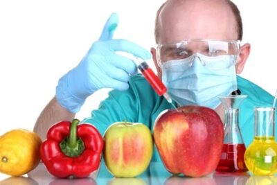 Scientist Injecting Apple in Lab Setting - genetiskt modifierade 9 Produce Test shows a masked, gloved researcher injecting a red apple with a syringe among other fruits and laboratory glassware.