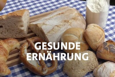 Assorted loaves, rolls, croissant, pretzel and a jar of flour arranged on a blue checkered cloth with the words gesunde ernahrung 1