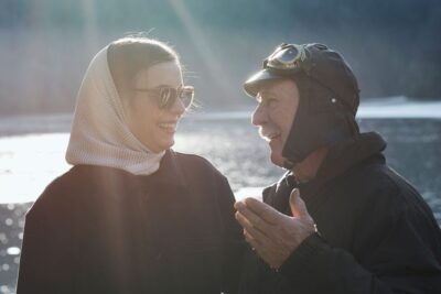 An older man in a vintage driving cap talks and smiles with a young woman in sunglasses and a headscarf by a sunlit lake, suggesting giovani conducenti and mentorship