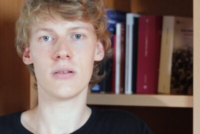 Close-up portrait of a blond young person in front of a bookshelf suitable for gli empiristi topics.