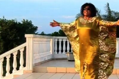 Person in a flowing gold robe with outstretched arms on a tiled terrace at sunset, grand pretre aesthetic.