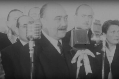 Black-and-white photo of a suited speaker at microphones during a formal press event about gratuidad universitaria