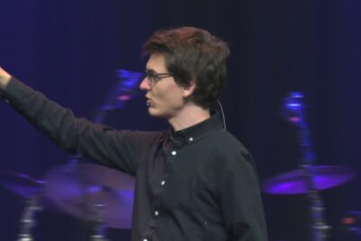 Side-profile of a man wearing glasses and a headset microphone speaking on stage with drums in the background during guds eksistens 6