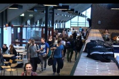 Crowded school commons with groups of students walking and carrying bags, gymnastikundervisning gymnasiet 1.