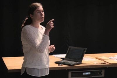 A woman standing beside a laptop and notes pointing while presenting about haitallinen ruoka and food safety