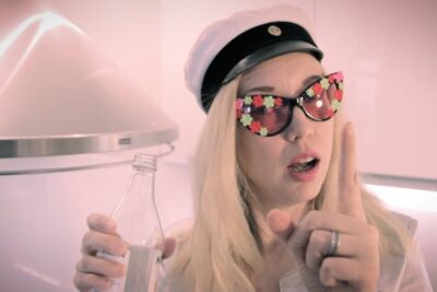 Person wearing a white sailor-style cap and flower-adorned sunglasses holding a bottle and pointing during halloweenin juhlimista.