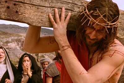 A man portraying holdning jesus carrying a wooden cross with a crown of thorns and visible wounds.