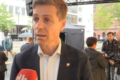 Man in dark suit speaking into a red microphone at an outdoor public event about homofilt ekteskap 6.