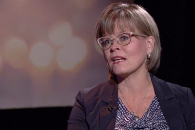 Middle-aged woman in a studio interview wearing glasses and a blazer discussing homoparien avioliitto oikeutta.