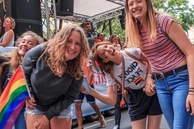 A group of smiling young people at a Pride parade holding a rainbow flag, celebrating homoseksuelle agteskaber 1.
