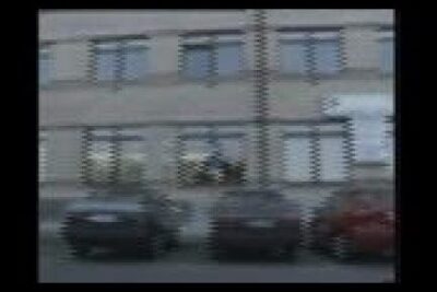 Blurry street scene of a multi-windowed building and parked cars suggesting themes of human cloning.