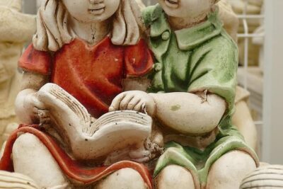 Two hand-painted ceramic children reading a book on a bench with worn patina, huolellisesti ja detailed