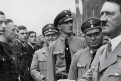 Black-and-white photo of a stern speaker and lined uniformed men at a public rally, imod krig