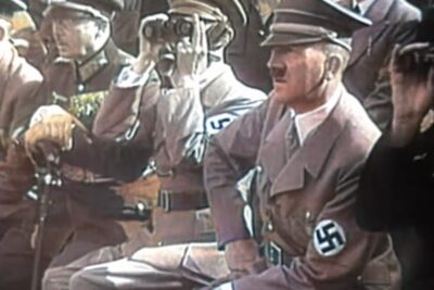 Seated uniformed leader in a brown coat with a visible swastika armband observing a public rally, imod parot doktrinen