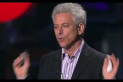 Gray-haired male speaker in a dark suit gesturing on stage, imot kolonialisme