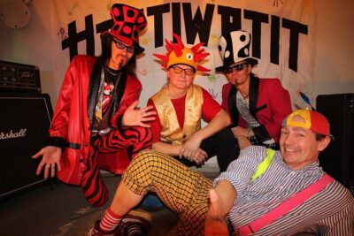 Four costumed performers in vibrant, mismatched outfits and novelty hats posing cheerfully onstage beneath a banner that references ja moraaliset.