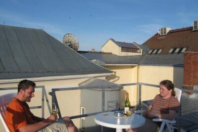 Two people seated on a sunlit rooftop terrace with a small round table, wine bottle and glasses ja vastatuuleen