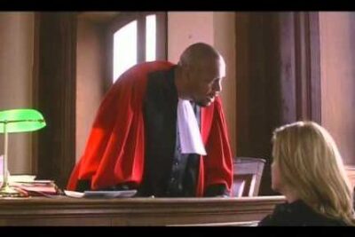A judge in a red robe leans over the bench to address a seated woman in a tense courtroom scene, jugement socrate.