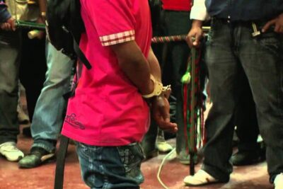 Person in a pink shirt with hands cuffed standing among a crowd of onlookers, justice indigene 1.