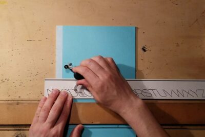 Hands tracing and cutting alphabet stencil on blue paper using a compass-like tool labeled k nstler 2