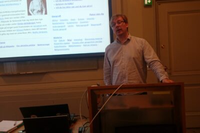 Person standing at a lectern giving a presentation with a projected webpage on the screen, kan vara 7.