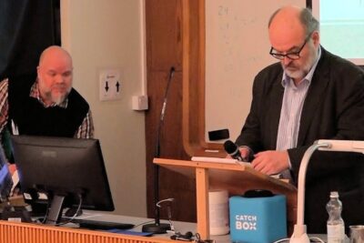 Speaker at a podium in a lecture hall presenting kirjallisen teoksen 1 with a second person at a computer