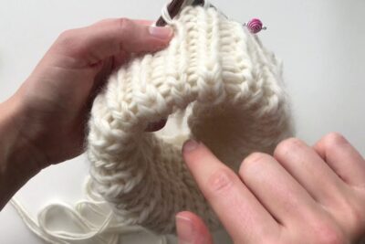 Hands knitting a cream chunky ribbed brim with stitch marker, krave samme