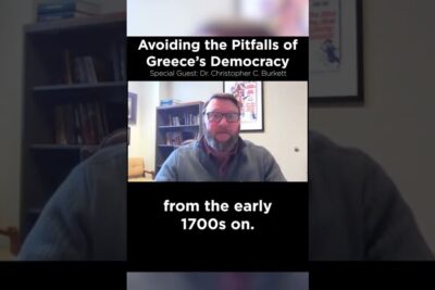 Video still of a seated speaker in an office with on-screen text about kreikan demokratian 1 and Greece's democracy.