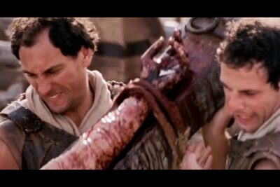 Two men in period garments strain to carry a blood-streaked wooden cross kreuzz ge 1