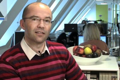 Man in striped sweater at office desk with bowl of fruit and monitors, suitable for kunstig befrugtning 1
