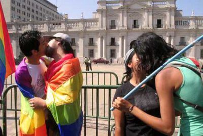 Two couples kissing wrapped in rainbow flags outside a stately government building, a public display addressing la discriminacion.