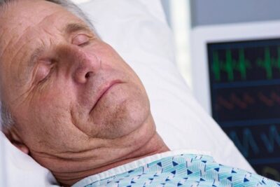 Older man with closed eyes lying in a hospital bed next to a cardiac monitor, la eutanasia 11