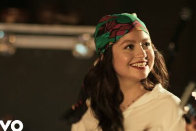 Smiling female musician wearing a colorful headscarf on stage with a guitar, la musica 1