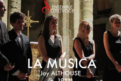 Choir members in black dresses and suits standing in a stone chapel beneath signage reading la musica 2