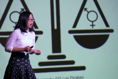 Presenter speaking in front of a projected balance scale with gender symbols illustrating langue inclusive 1.