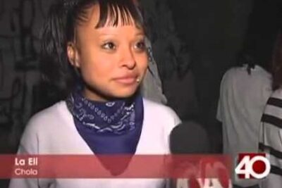 Person with short bangs and a blue bandana being interviewed against a graffiti wall, styled for las gafas 1 eyewear visuals.