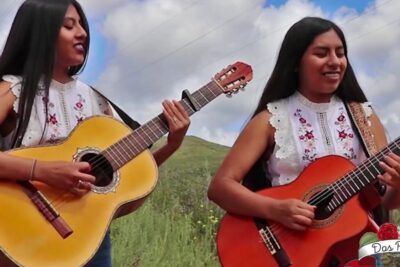 Two women playing acoustic guitars outdoors wearing embroidered floral tops featuring las rosas motif.