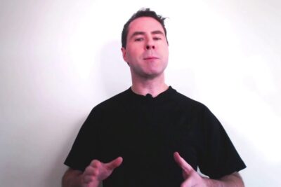 Man wearing a black t-shirt and lavalier microphone gesturing in front of a white wall about lassurance universelle.