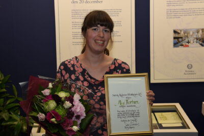 Smiling woman holding a framed certificate and bouquet at an award presentation leda till 4