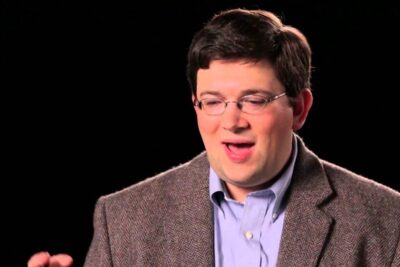 Portrait of a man in a tweed jacket and glasses speaking about legal precedents against a dark background.