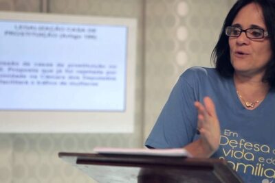 Woman at a lectern gesturing while presenting with a blurred projection screen, wearing a blue T-shirt and glasses, legalizacao prostituicao 1
