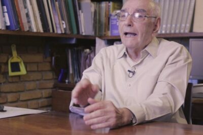 Senior academic seated at a wooden desk with bookshelves behind him speaking about lei universitaria