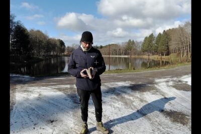 Person in a dark jacket and beanie holding a book by a partially frozen lake lessere umano 1