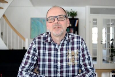 Man wearing glasses and a purple plaid shirt seated in a bright home office, levende organisme 2