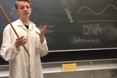 Person in a lab coat pointing at a chalkboard with DNA diagrams for levende organismer 1.