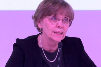 Female conference speaker wearing glasses and a pearl necklace speaks into a microphone about lexperimentation humaine.