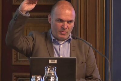 A man speaking at a lectern with laptop and microphone during a formal presentation, liberaali kauppapolitiikka 1